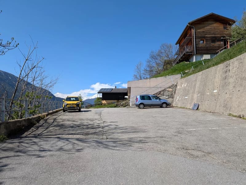 The image shows a rural mountain setting with a wooden chalet-style building on a hillside. There is a paved parking area in the foreground with two cars parked, one yellow and one gray. The building has a balcony and appears to be surrounded by trees and
