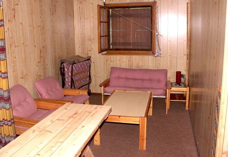 Wooden cabin interior with a living room area, including a sofa, coffee table, and a small table. There is a window with a wooden frame visible on the wall.