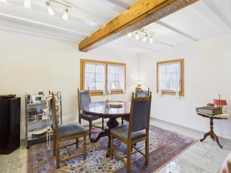 Spacious dining room with wooden beams on the ceiling, a round wooden dining table with chairs, a rug on the floor, and a fireplace visible in the room.