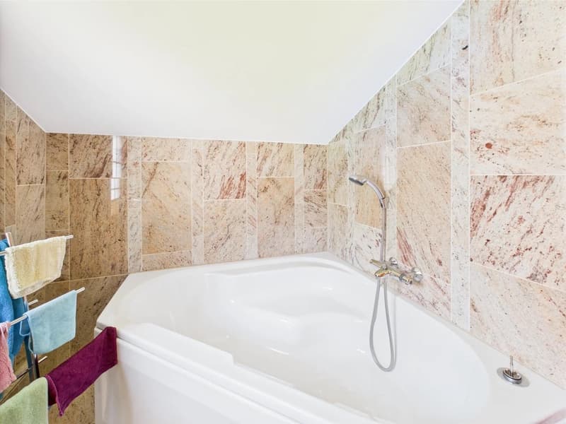 Spacious bathroom with beige marble-like tiles covering the walls, a white bathtub, and a shower fixture with a handheld showerhead. Towel racks are visible on the side.