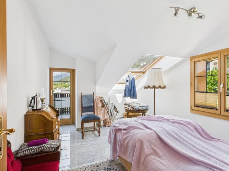 Spacious attic bedroom with slanted ceilings, large windows providing natural light and a view, wooden furniture including a dresser and chair, a bed with pink bedding, and a lamp on a side table.
