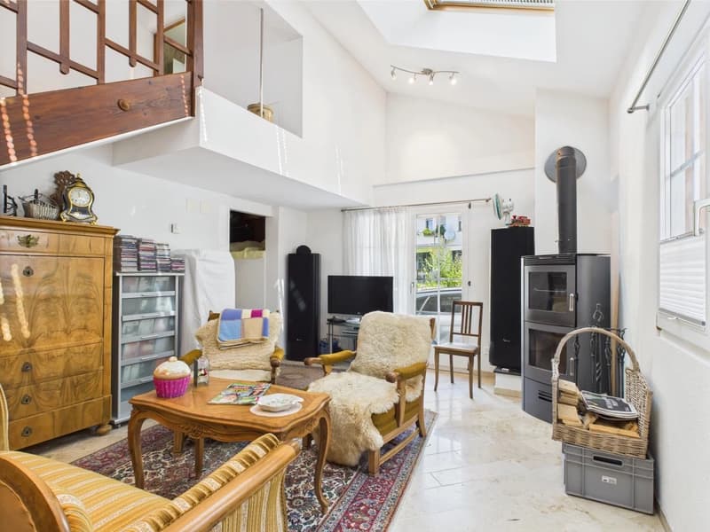 Spacious living room with high ceilings, wooden beams, and large windows. Furnished with a wooden dresser, armchairs, a coffee table, and a TV stand. The room also features a fireplace and a tiled floor.