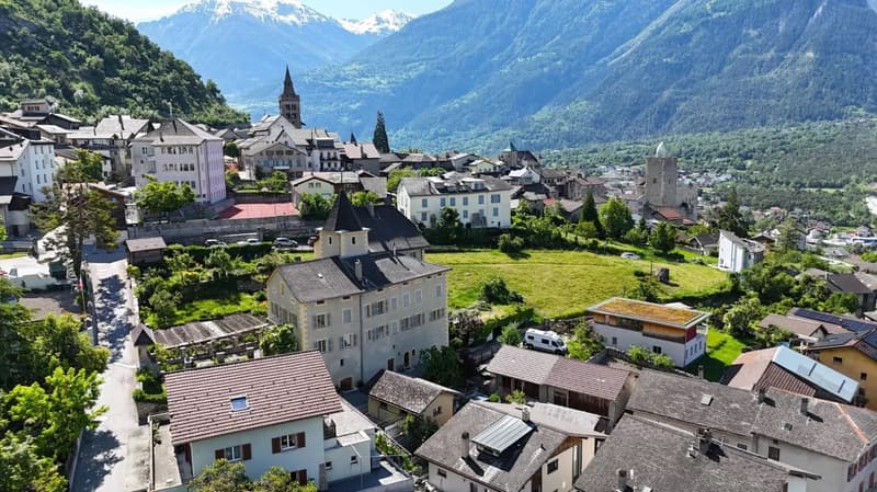 This image shows a picturesque town nestled in a valley surrounded by mountains. The town features a mix of traditional European architecture, including churches with tall spires, as well as residential and commercial buildings. The town is situated on a