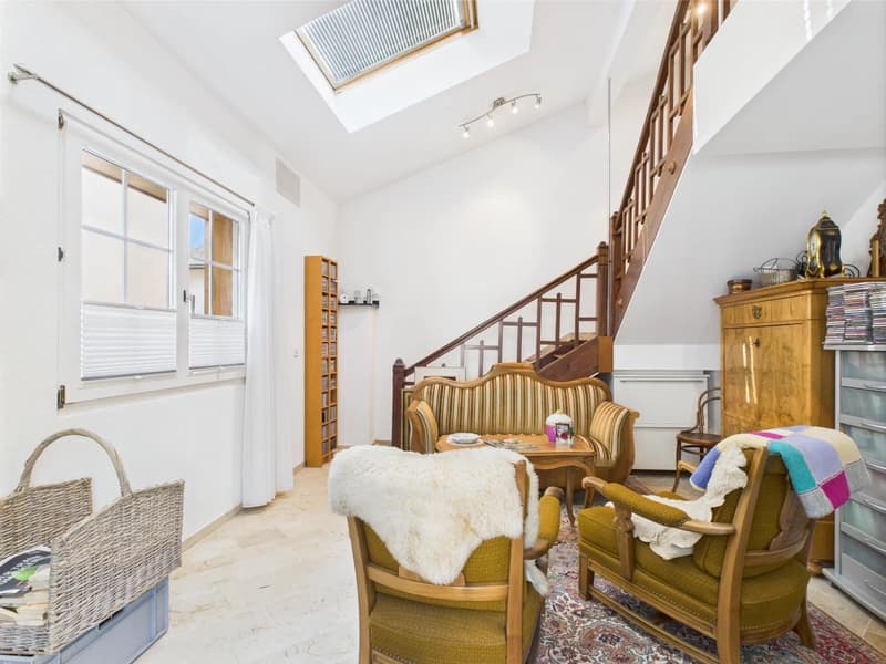 Bright and airy living room with a vaulted ceiling, skylights, and large windows. The room features a wooden staircase, a vintage-style sofa and armchair, a wooden bookshelf, and a decorative cabinet. The space has a cozy and eclectic feel with the additi