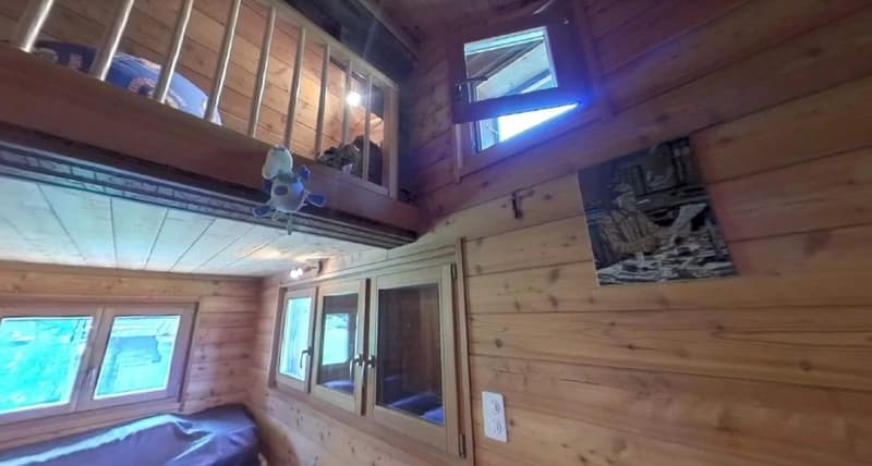 Wooden cabin interior with large windows, loft area, and decorative elements such as a stuffed animal and artwork on the walls.