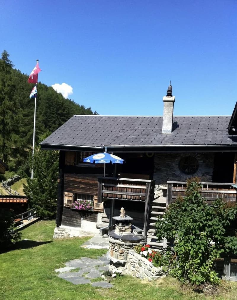 Traditional alpine-style wooden house with a slate roof, surrounded by trees and a grassy yard. There is a balcony or terrace on the front of the house, and a small fountain or water feature in the yard.