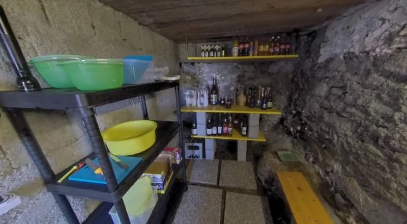 Underground storage room with concrete walls, shelving units holding various containers and bottles, likely used for storage of household items or supplies.