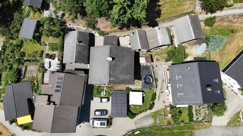The image shows an aerial view of a residential area with several houses and buildings. The roofs of the buildings are mostly dark-colored, and there are some cars parked on the property. The surrounding area appears to be mostly green, with trees and veg