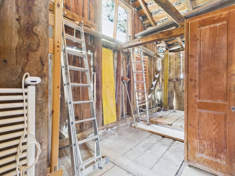 The image shows the interior of a partially renovated or under-construction building. The walls are made of wooden beams and panels, and there is a ladder and various construction materials visible. The floor appears to be made of wooden planks. The overa
