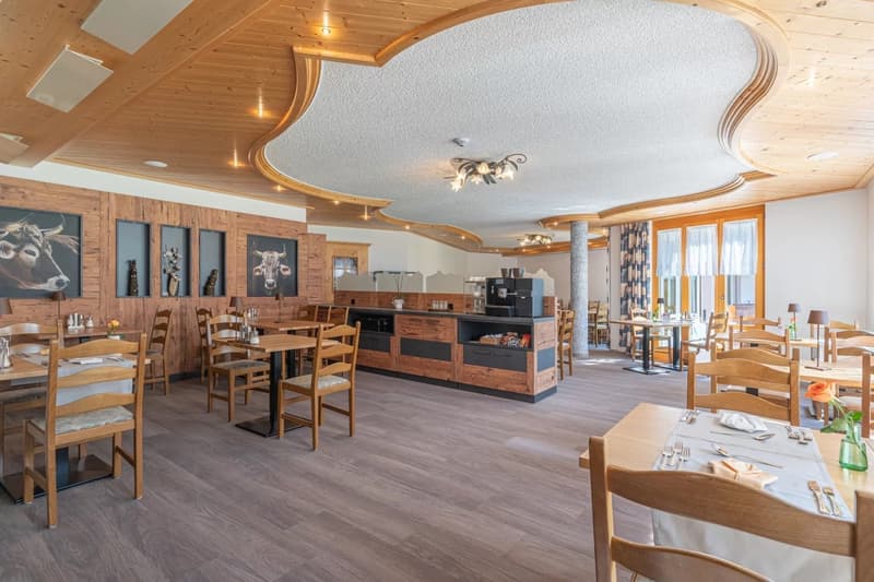 Large open dining area with wooden walls and ceiling, wooden tables and chairs, a kitchen counter with appliances, and decorative cow head wall art. The room has a warm, rustic feel with a fireplace visible.