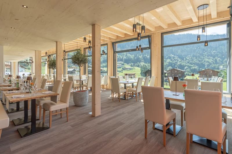 Spacious dining area with large windows offering scenic mountain views, wooden beams and columns, wooden tables and chairs, pendant lighting fixtures, and a cozy, rustic atmosphere.