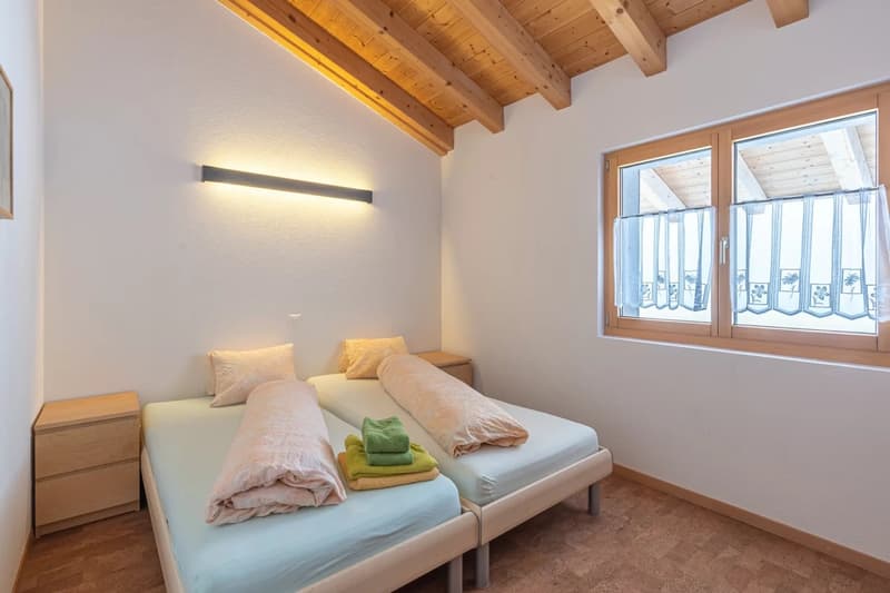Spacious bedroom with wooden beamed ceiling, large windows, two twin beds, bedside tables, and a wall-mounted light fixture.