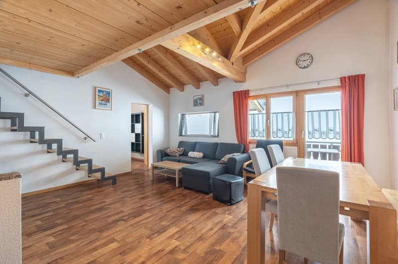 Open-concept living room with wooden ceiling beams, hardwood flooring, large windows, a sofa, coffee table, and dining table
