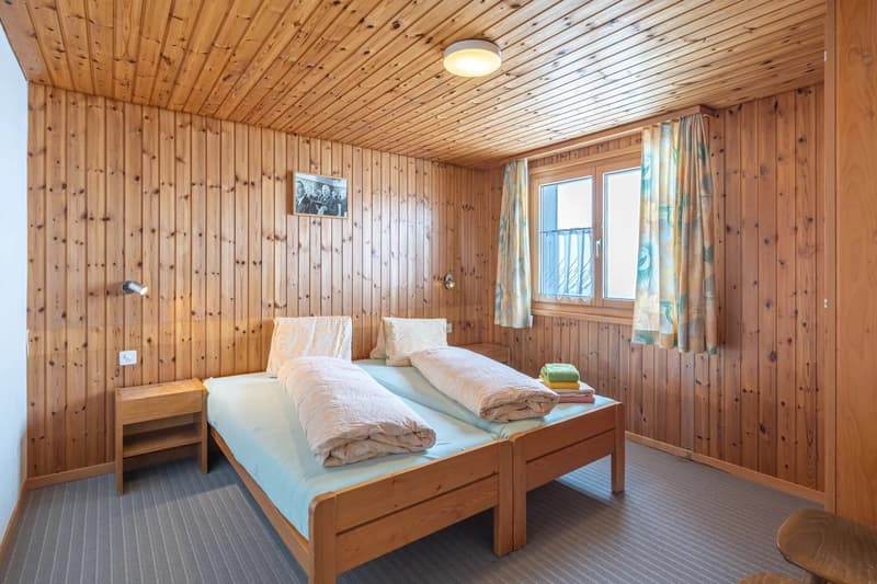 Cozy wooden bedroom with two twin beds, wooden walls and ceiling, large window, curtains, and a small side table