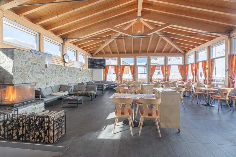 Large open-concept dining area with wooden beamed ceiling, stone fireplace, comfortable seating, and wooden tables and chairs. The space has large windows providing a scenic view of the snowy outdoor landscape.