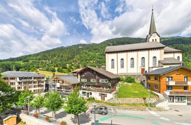 Picturesque alpine town with a church steeple, traditional wooden buildings, and a parking area in the foreground. The town is surrounded by lush green hills and forests.
