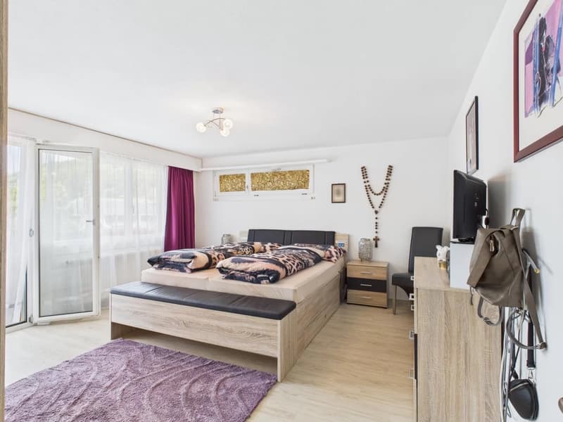 Spacious bedroom with large windows, wooden flooring, a king-size bed with a patterned comforter, bedside tables, a dresser, and a TV mounted on the wall. The room also features a purple curtain and a decorative wall hanging.