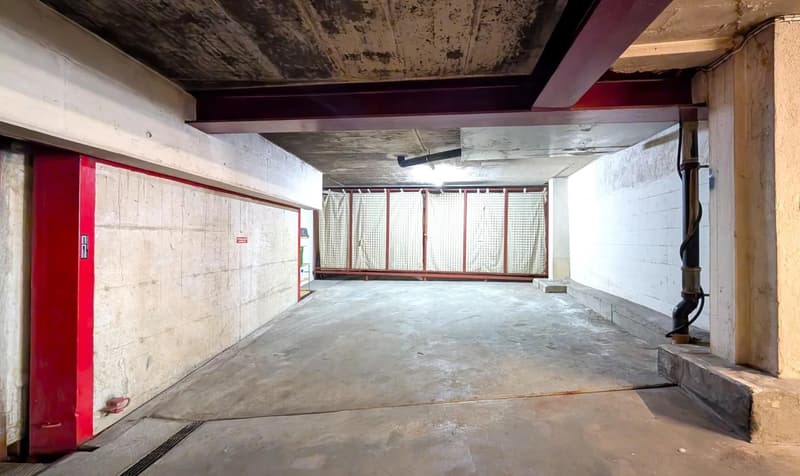 Large open garage space with concrete floor, exposed ceiling beams, and a red metal door.