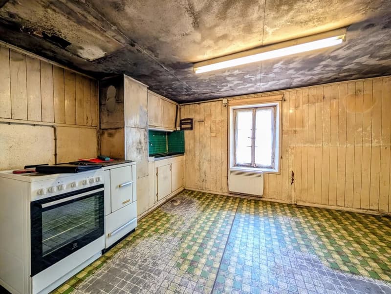 Dilapidated kitchen with old wooden walls, damaged ceiling, and a tiled floor in a mix of blue and green colors. The room contains an old stove and some cabinets.