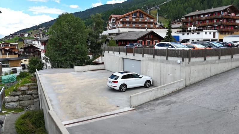 The image shows a residential area in a mountainous region, with several multi-story buildings and chalets. There is a parking area in the foreground with a white car parked. The buildings have a traditional alpine architectural style with wooden elements
