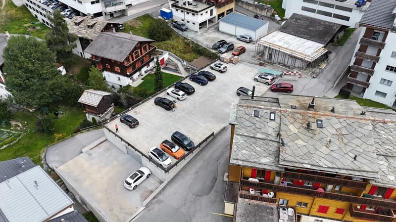 The image shows an aerial view of a commercial and residential area. There are several multi-story buildings, including apartment buildings and commercial structures. There is a paved parking area with various cars parked. The surrounding area includes so