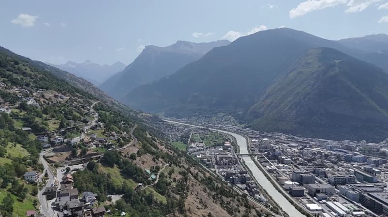 Aerial view of a valley with a winding road, a town, and a river flowing through it, surrounded by mountains with some snowy peaks.