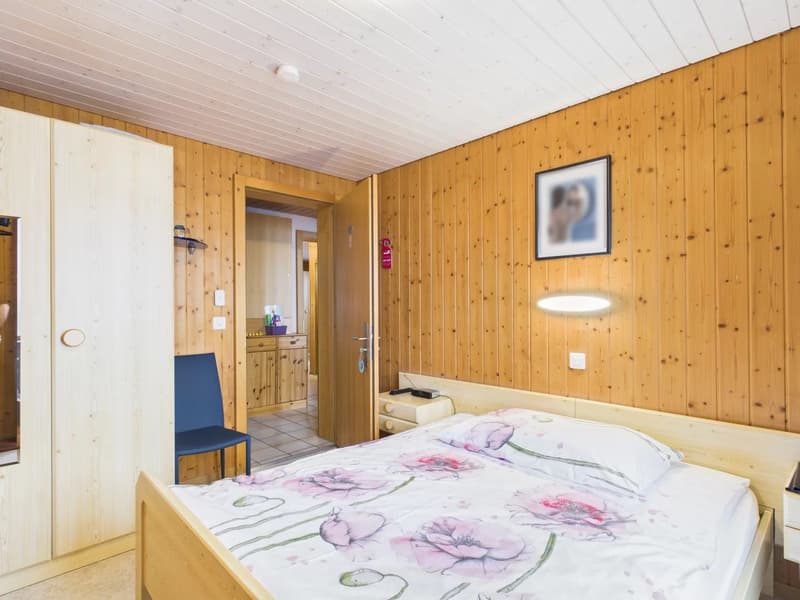 Bedroom with wooden walls, bed with floral bedding, chair, bedside tables, wall picture, and open door to the hallway.