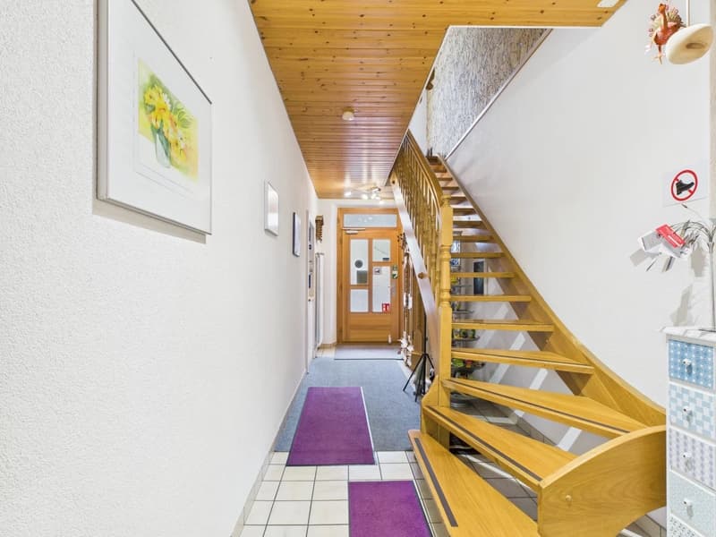 Entrance hallway with white walls and tiles, wooden spiral staircase, two purple floor rugs, picture frame, light fixtures, and door at the end