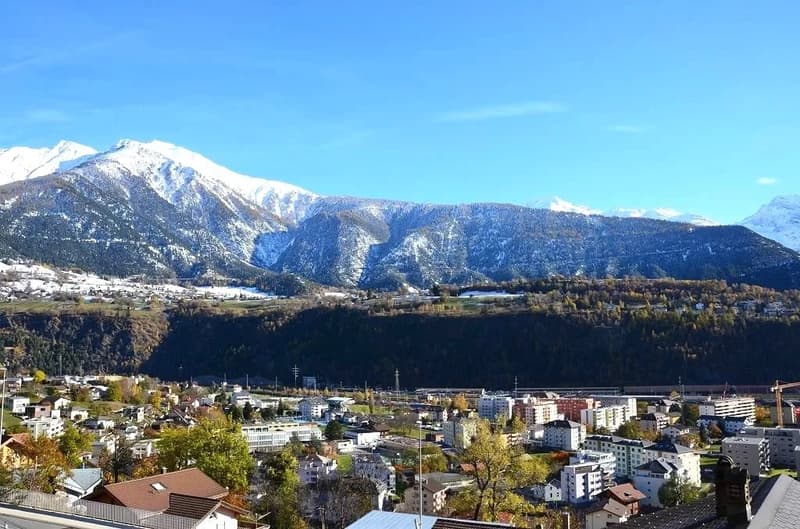 View of mountains with snow in background, surrounded by a town with various buildings, trees and greenery