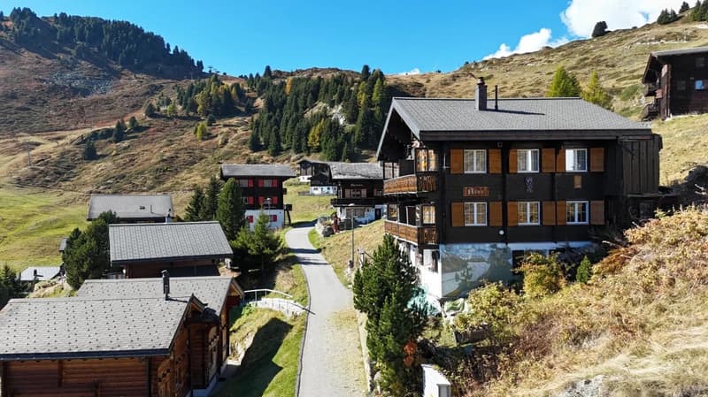 A group of wooden chalets, with grey roofs, set on a hill overlooking a valley. There are trees and grass on the hill. One of the chalets has a balcony with wooden railings.
