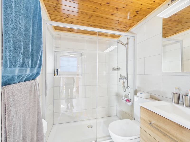 A modern bathroom with glass shower, white tiles, wooden ceiling, wooden cabinet with sink, toilet, and a mirror. Towels and toiletries are on the towel rack.