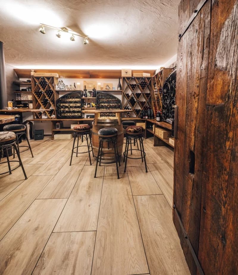 Wine cellar with wooden flooring, wooden door, wine racks, wooden barrel table, stools, wine bottles, wooden shelves, lighting fixtures