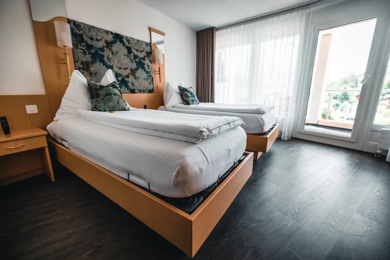 Two beds with white sheets, wooden headboards, nightstand with remote, mirror, windows with curtains, balcony
