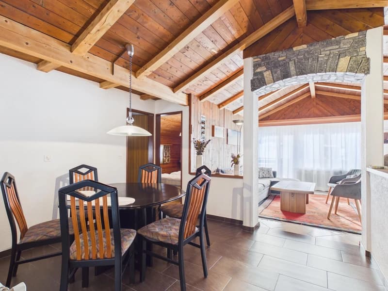 Dining room with wooden table and chairs, wooden ceiling, white walls, wooden floor, lamp, couch, coffee table, rug, and a window with curtains.