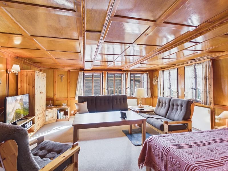 living room with wooden walls, sofa, coffee table, TV, lamp, bed, bedspread, carpet, wooden chair, wooden cabinet, shelves, books, cross, windows, curtains