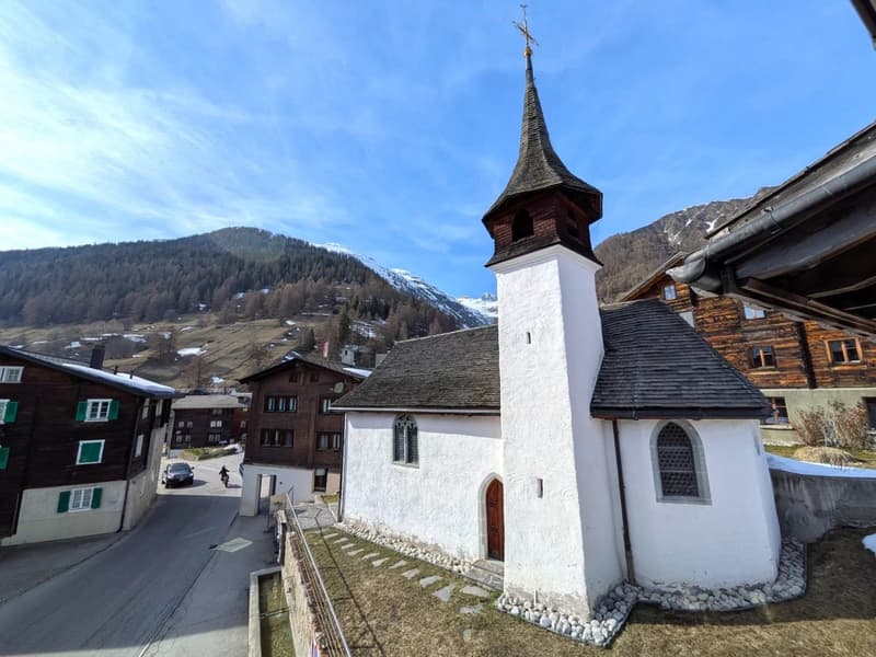 small church with a steeple, wooden door, windows, surrounded by houses, mountains in the background