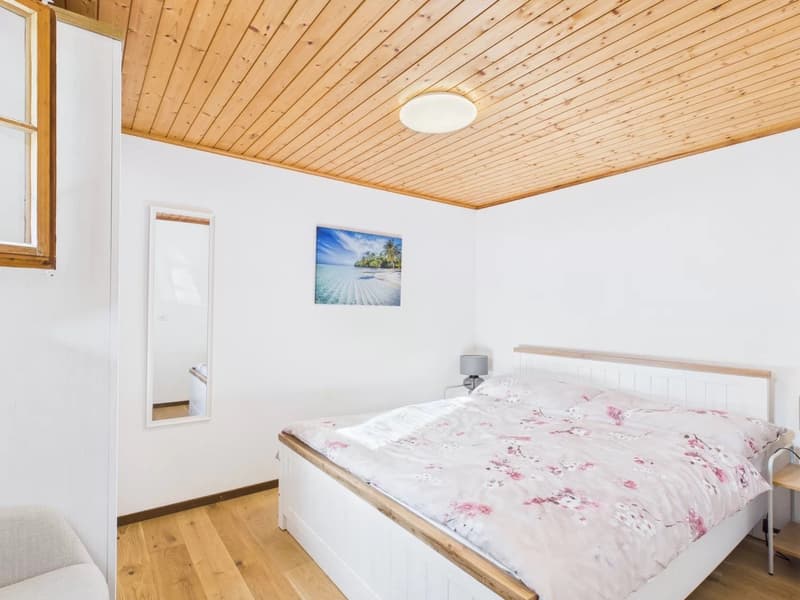 Bedroom with wooden floor, white walls, wooden ceiling, bed, nightstand, lamp, mirror, picture frame, and a chair.