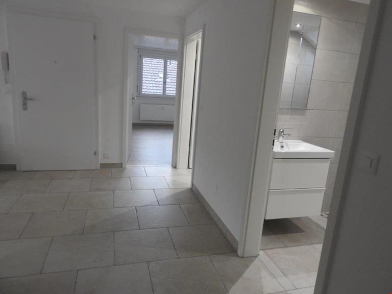 Empty room with white walls, tiled floor, white door, white sink, mirror, and radiator