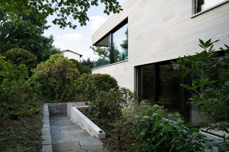 Modern house with large glass windows, concrete walls, pathway leading to entrance, garden with various plants