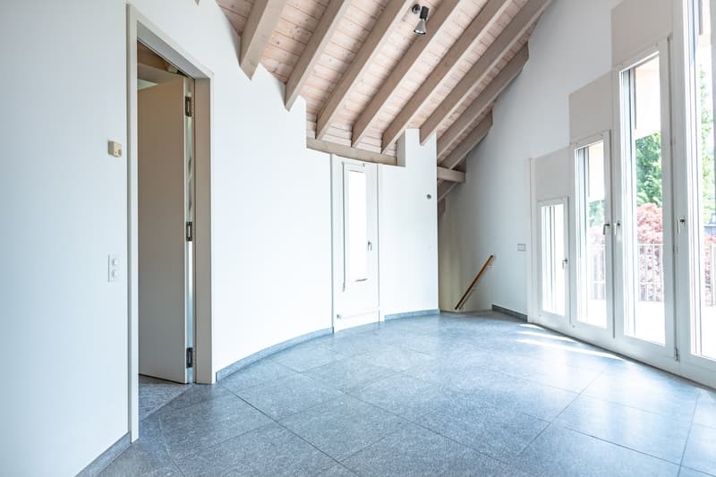 Spacious room with high vaulted ceiling, wooden beams, and large windows providing natural light. The room has a tiled floor and a sliding glass door leading to a balcony or terrace.