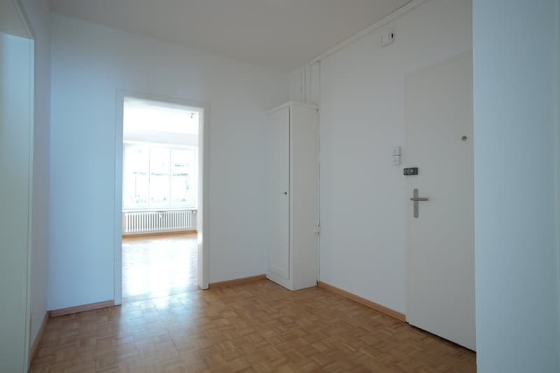 An empty room with white walls and wooden floors. There is a white door, a white wardrobe, and windows.