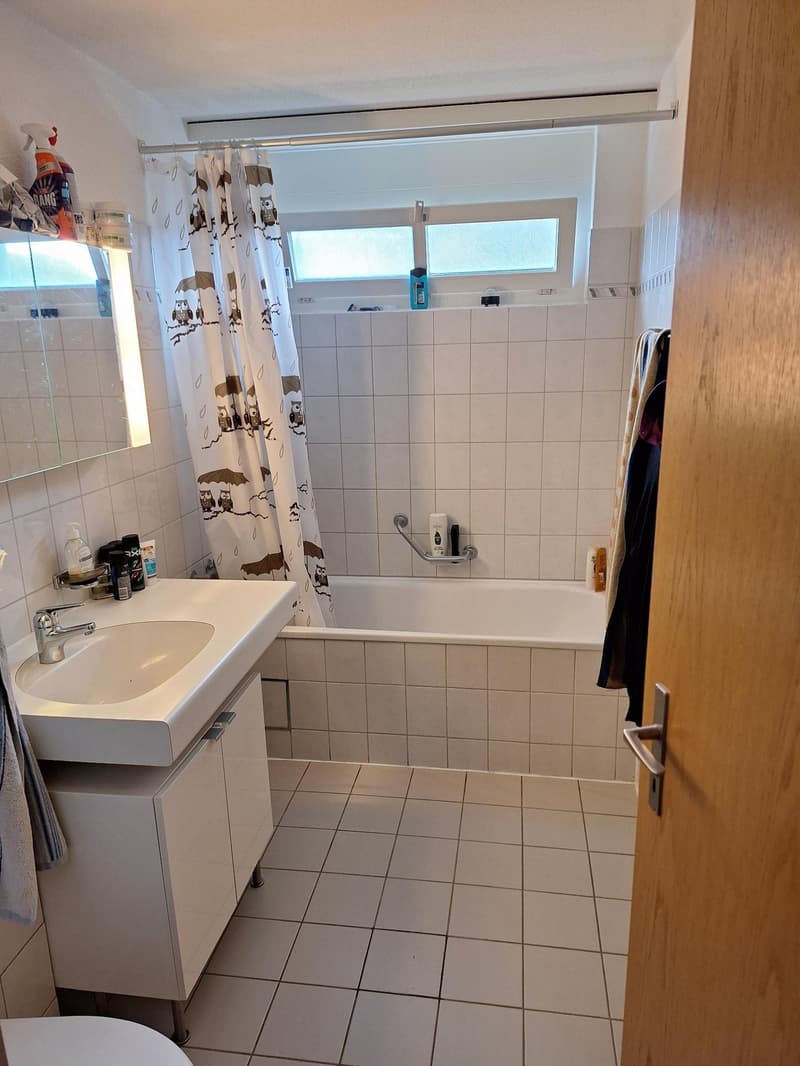 Bathroom with white tiles, bathtub, sink, mirror, wooden door, towel rack, and various toiletries