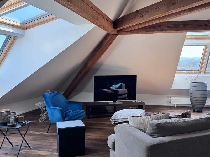 Spacious attic living room with exposed wooden beams, large windows providing natural light, blue velvet armchair, gray sofa, TV stand, and decorative vase