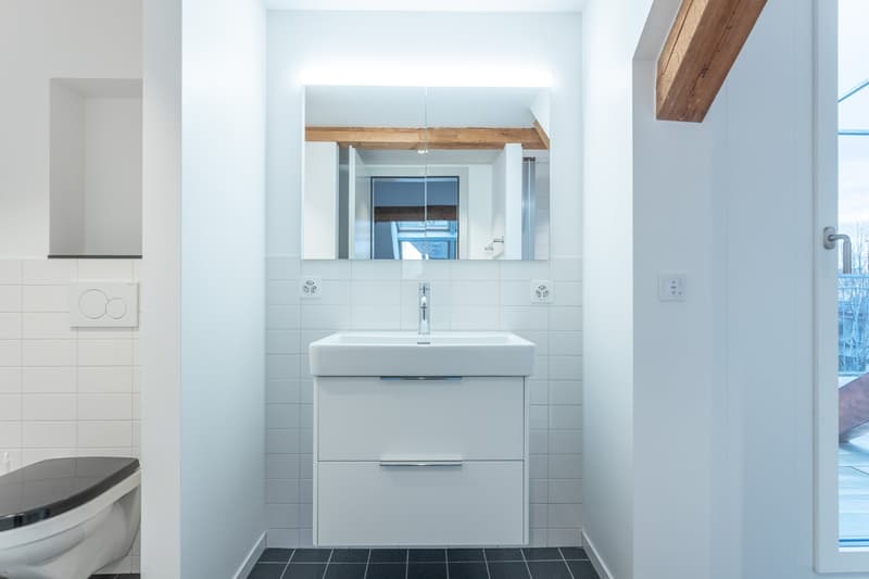 Modern bathroom with white tiles, a white vanity unit with a sink, and a large mirror above it. The room has exposed wooden beams on the ceiling and a window providing natural light.