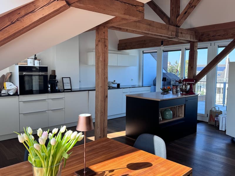 Spacious kitchen with exposed wooden beams, modern white cabinets, black kitchen island, hardwood flooring, and large windows providing natural light and a view of the balcony or terrace.