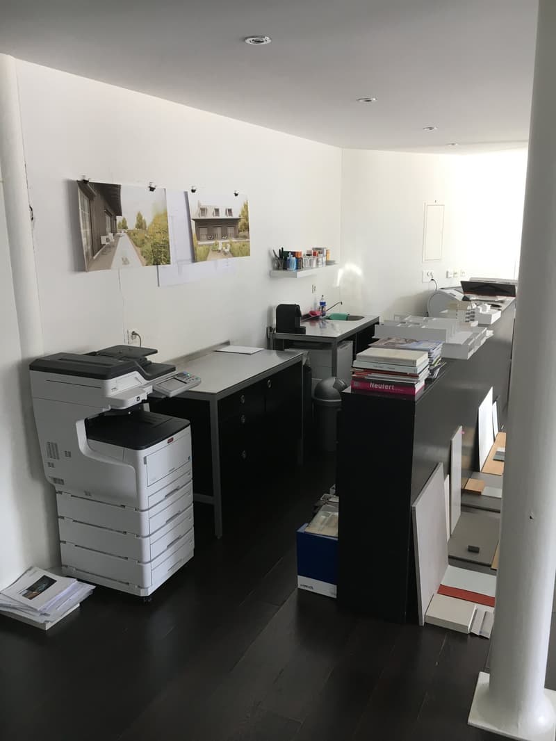 The image shows an office space with various office equipment and supplies. There are two printers/copiers, a desk with a computer, and shelves with books and other items. The walls are decorated with artwork. The overall impression is of a functional and