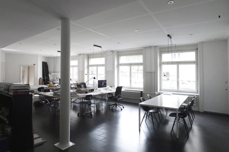 Open office space with large windows, desks, chairs, and computer equipment. The room has a minimalist, modern design with a neutral color palette.