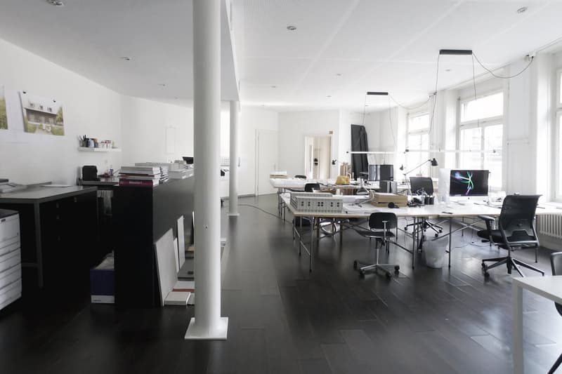Large open office space with multiple desks, computers, and office equipment. The room has high ceilings, white walls, and dark hardwood floors. There is a dishwasher visible in the corner of the room.