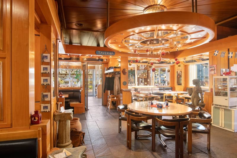 The image shows a cozy and rustic-style restaurant or taverna with wooden walls and ceilings, a large circular chandelier, round wooden tables and chairs, and various decorative items and artwork on the walls. The space appears to have a warm and inviting