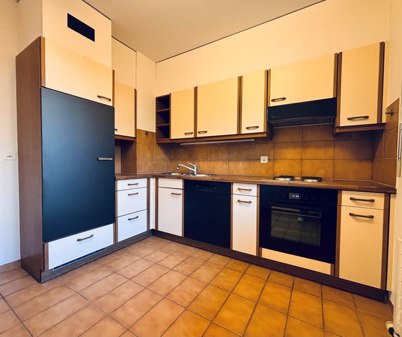 A modern kitchen with dark cabinets, white tiled floor, stainless steel sink, dishwasher, washing machine, gas stove, and overhead cabinets.